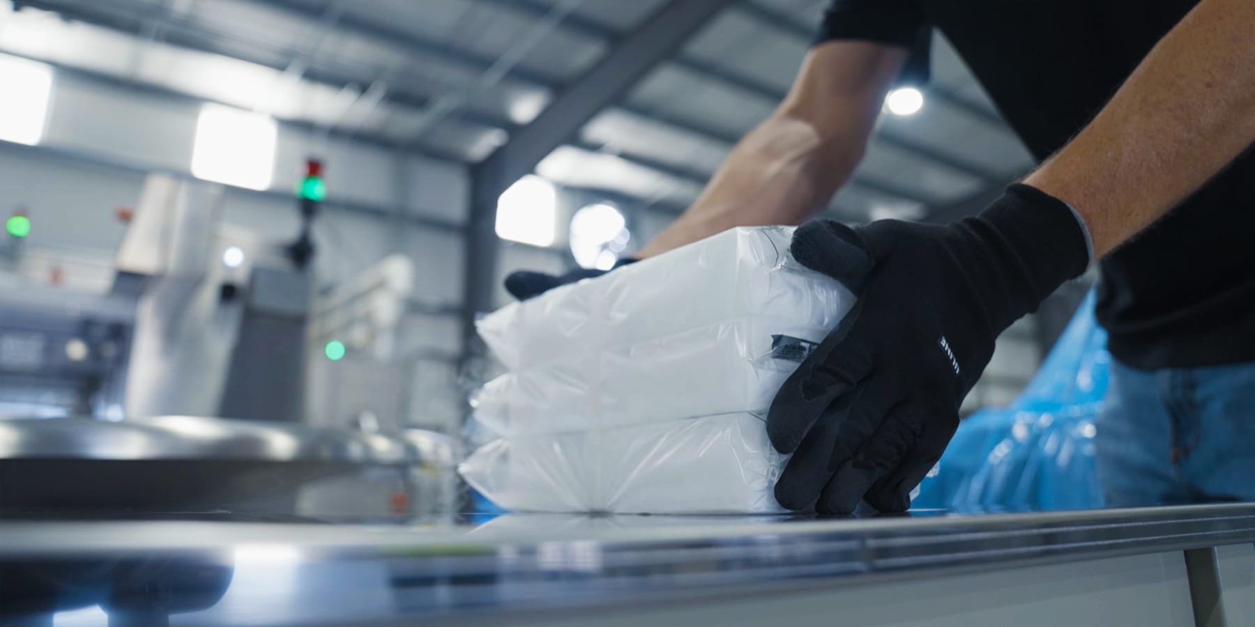 Ein Mann in dunkler Arbeitskleidung und schwarzen Handschuhen hantiert mit einer Gruppe von weißen, verpackten Trockeneisscheiben auf einer metallenen Arbeitsfläche. Der Hintergrund zeigt unscharfe industrielle Anlagen.