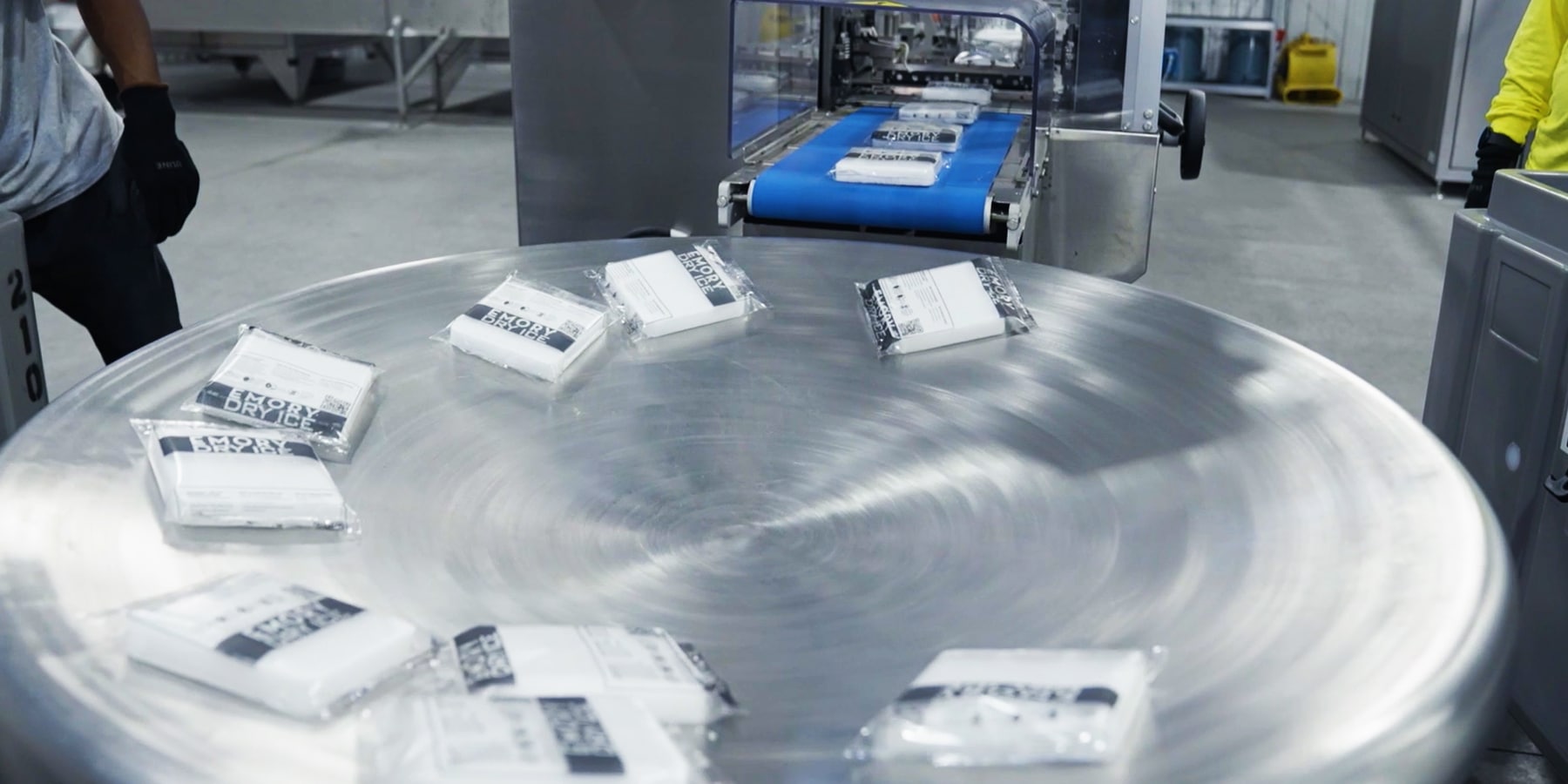 Eine Nahaufnahme eines runden, metallischen Tisches in einer Industrieumgebung. Mehrere weiße, verpackte Trockeneisscheiben sind unregelmäßig darauf angeordnet. Im Hintergrund ist eine Förderbandanlage und weitere industrielle Maschinen zu sehen.