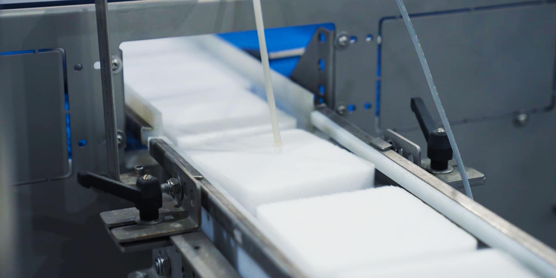 Eine Nahaufnahme einer industriellen Maschine, die rechteckige, weiße Trockeneisscheiben auf einem Förderband verarbeitet. Die Blöcke bewegen sich durch eine Vorrichtung mit Metallteilen und Schrauben.