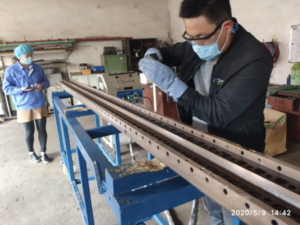 Na zdjęciu przedstawiono mężczyznę w maseczce ochronnej, okularach i rękawicach, który trzyma dyszę urządzenia do czyszczenia suchym lodem i prowadzi proces czyszczenia elementu metalowego. Wygląda to na głowicę lub matrycę z wieloma otworami, prawdopodobnie element linii do produkcji włókniny. W tle, po lewej stronie, stoi druga osoba w maseczce, czepku i fartuchu, trzymająca urządzenie mobilne lub notatnik — wygląda, jakby dokumentowała proces lub nadzorowała przebieg prac.