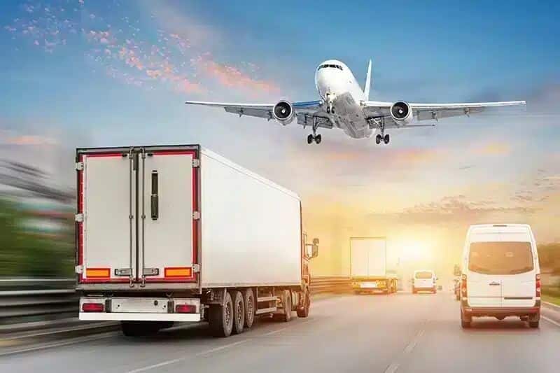 Na grafice przedstawiono dynamiczną scenę transportową, ukazującą współczesną logistykę w ruchu. Na pierwszym planie jedzie biały ciągnik siodłowy, towarzyszy mu dostawczy van i inne pojazdy na wielopasmowej autostradzie. Nad drogą przelatuje samolot pasażerski, uchwycony w momencie wznoszenia lub lądowania na tle nieba w barwach wschodu lub zachodu słońca.