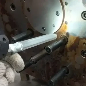 Obraz przedstawia zbliżenie mechanicznego układu, prawdopodobnie będącego częścią maszyny przemysłowej lub procesu produkcyjnego. Widać okrągły metalowy element z kilkoma otworami i śrubami, z którego wystają cztery cylindryczne pręty. Ubrana w rękawiczkę ręka trzyma maszynę do czyszczenia suchym lodem. 